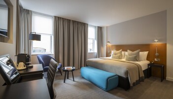 Hotellihuone, jossa on sänky, työpöytä, kahvinkeitin, tuoli, pieni pöytä ja sininen rahi.