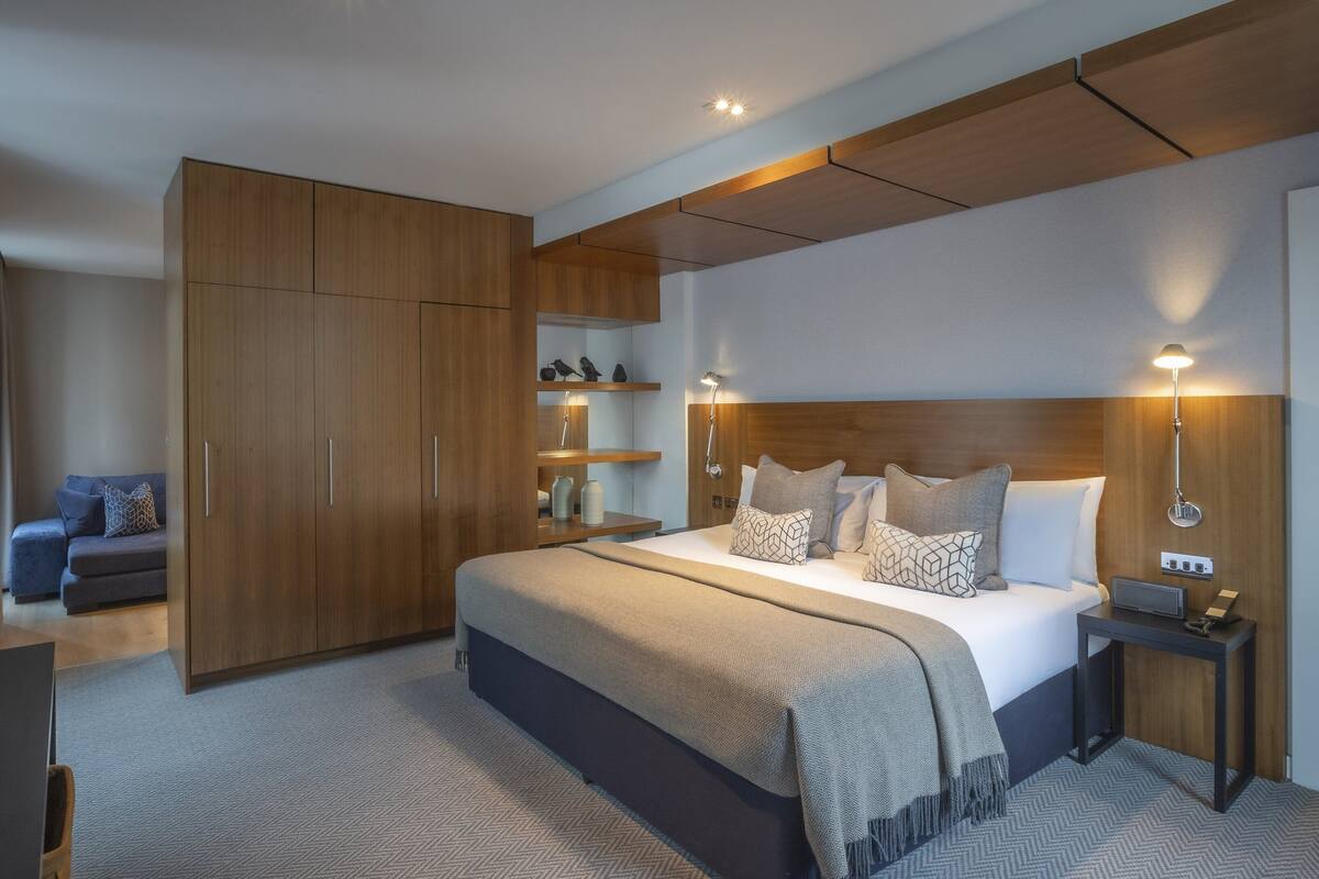 A modern hotel room with a large bed, wooden wardrobe, bedside tables, and a desk.