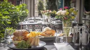 Een eettafel met een hamburger, friet en een visbroodje, omringd door glazen en een vaas met bloemen.