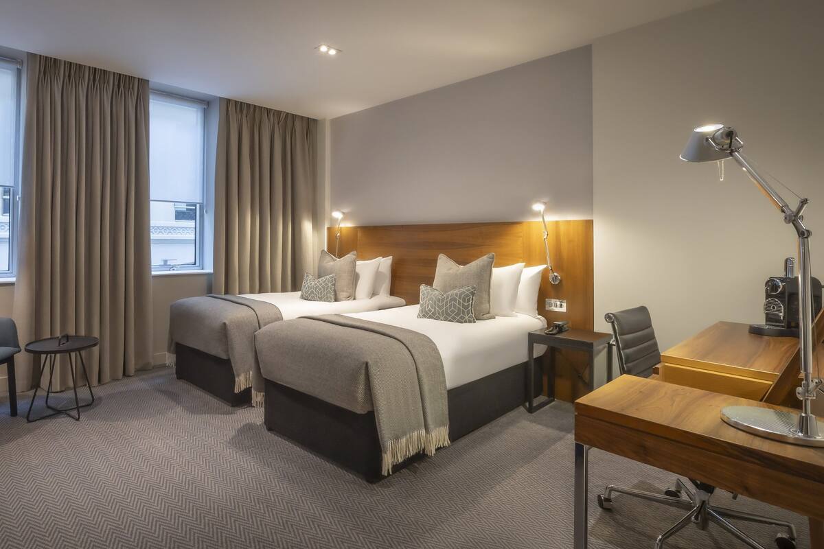 A hotel room with two beds, a desk, a chair, a TV, and a lamp.