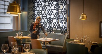 Een restaurantinterieur met een modern geometrisch wandontwerp, houten tafels en zitplaatsen met groene bekleding.