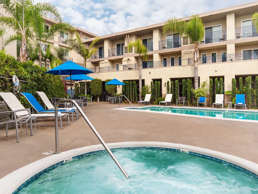 A hotel pool area with multiple lounge chairs and umbrellas, surrounded by palm trees and a multi-story building.