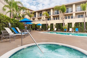 Zona de piscina de hotel con múltiples sillas de descanso y sombrillas, rodeada de palmeras y un edificio de varios pisos.