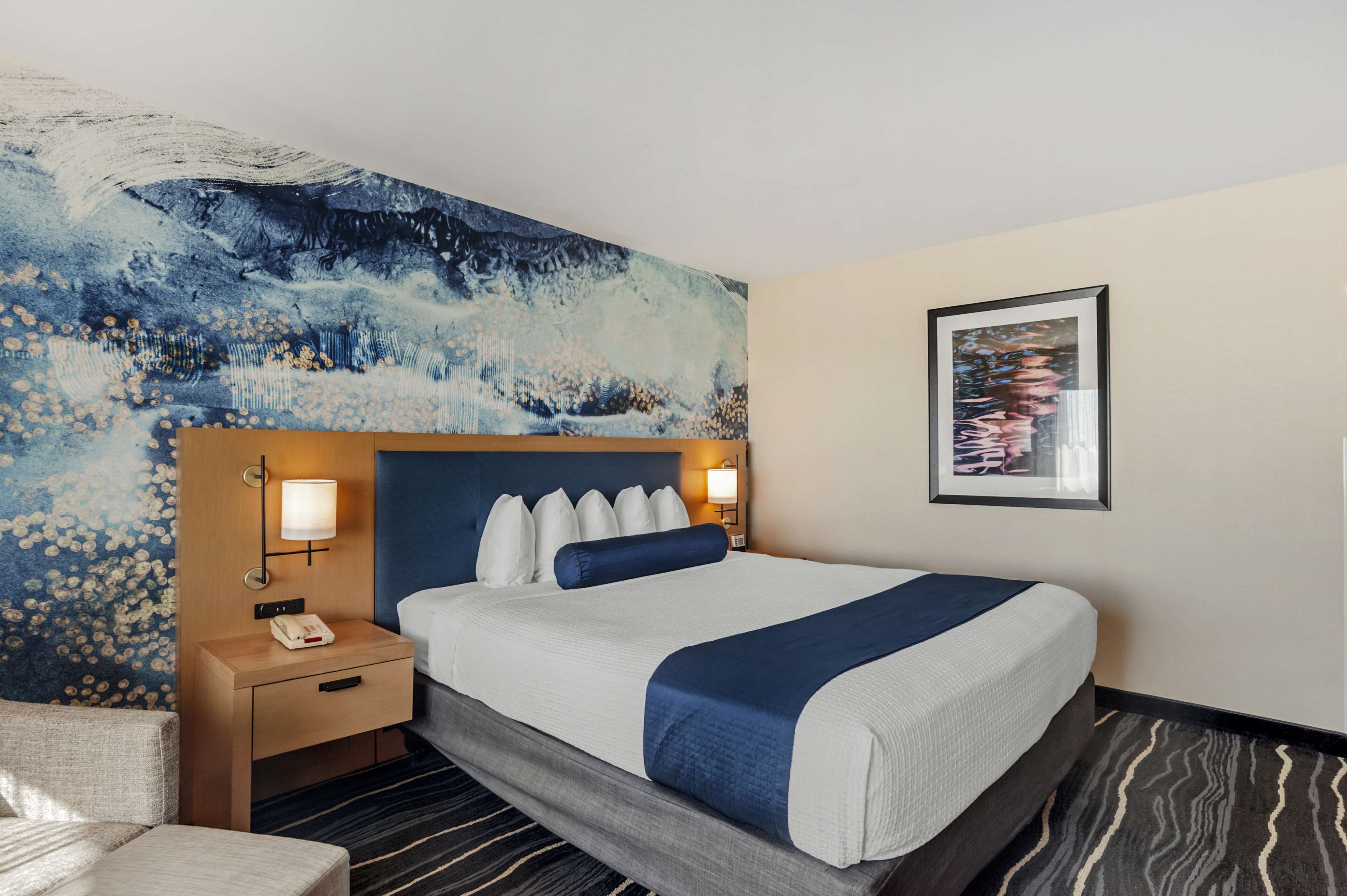A hotel room with a large bed, a nightstand with a phone, a wall with a painting, and a carpeted floor.