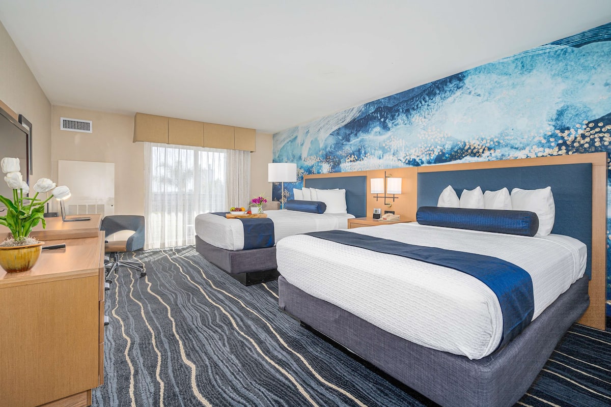 Habitación de hotel con dos camas, un escritorio, una silla, un ventanal con cortinas y un mural en la pared.