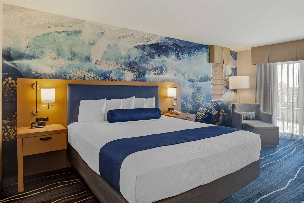 Habitación de hotel con una cama grande, dos mesitas de noche con lámparas, una silla y un amplio mural abstracto en la pared.
