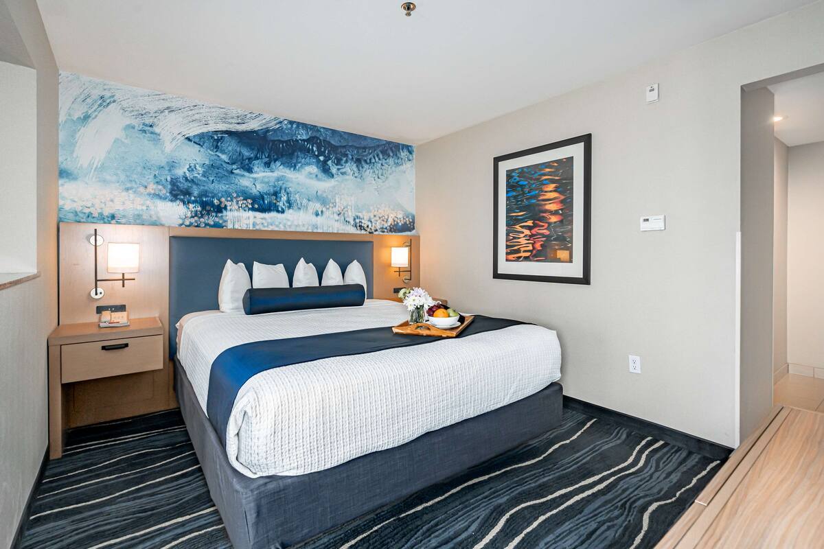 Una habitación de hotel con una cama grande, una mesita de noche, una lámpara fijada a la pared, una bandeja con frutas y flores, y un cuadro abstracto de gran tamaño sobre la cama.