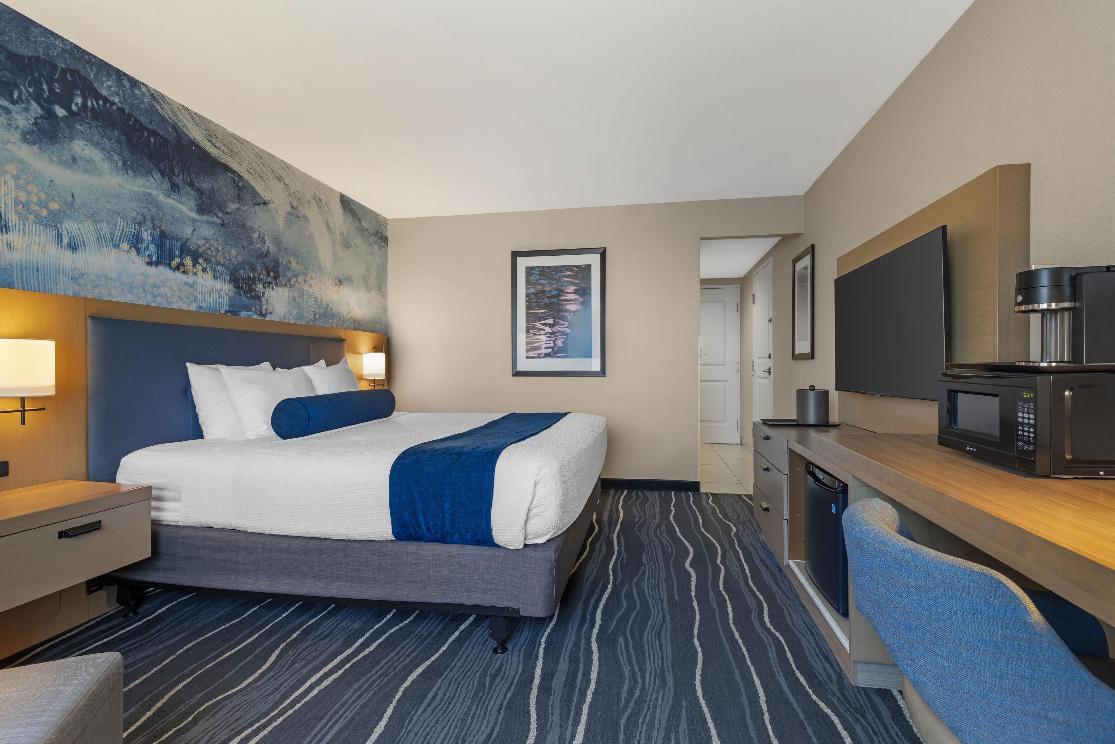 A hotel room with a bed, bedside table, lamp, artwork, television, and a microwave.