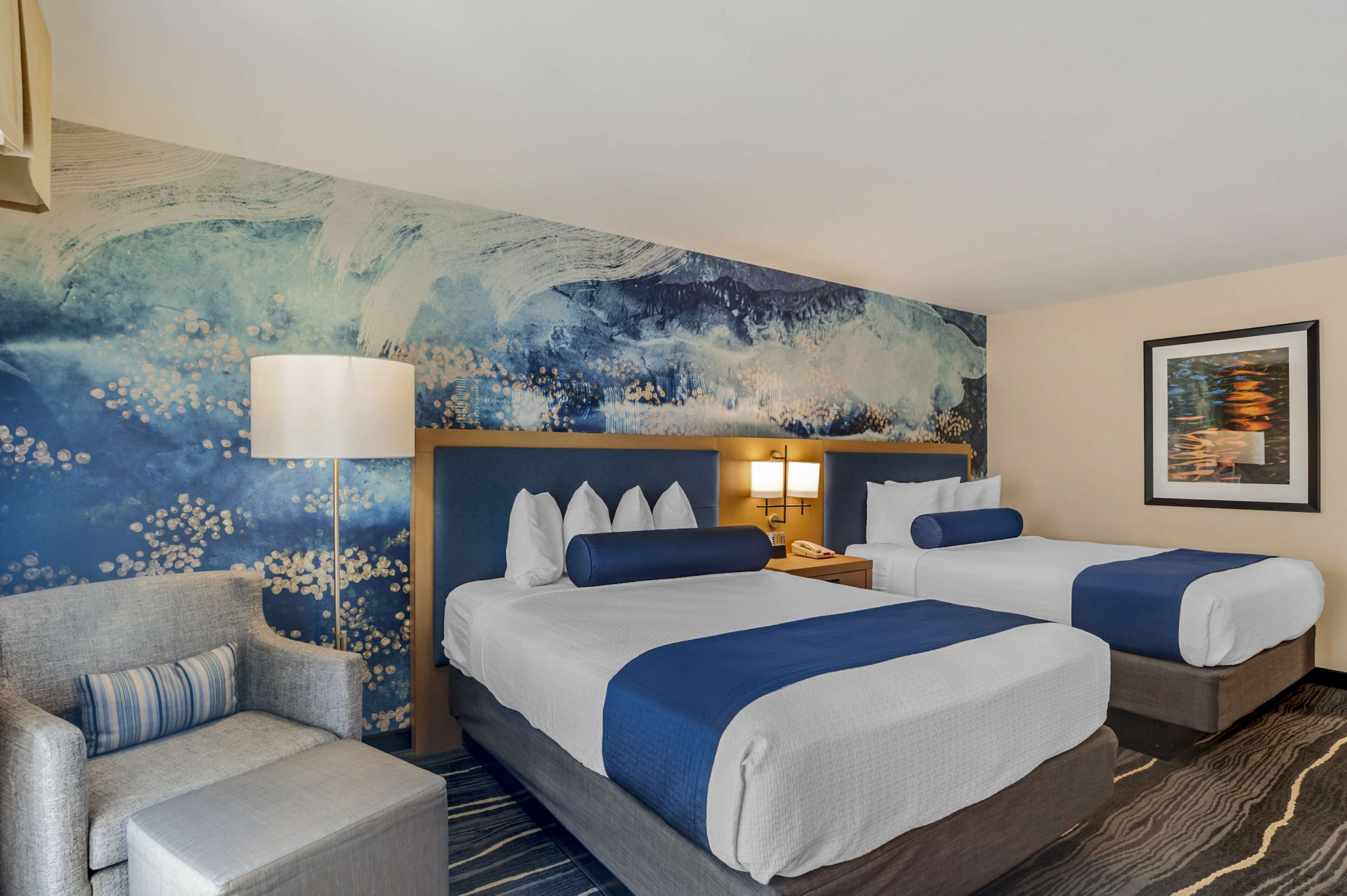 Habitación de hotel con dos camas, un sofá, un cuadro abstracto grande y una mesita de noche con lámpara.