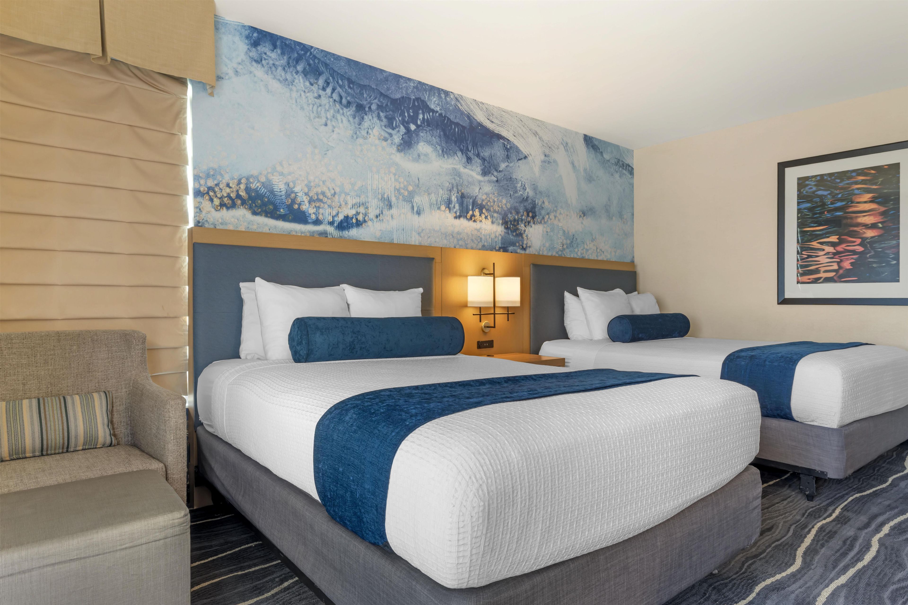 Een hotelkamer met twee bedden, een bank, een groot abstract schilderij en een lijst met een foto aan de muur.