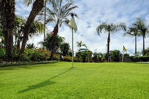 A well-maintained lawn with a flagpole, palm trees, and a building in the background.