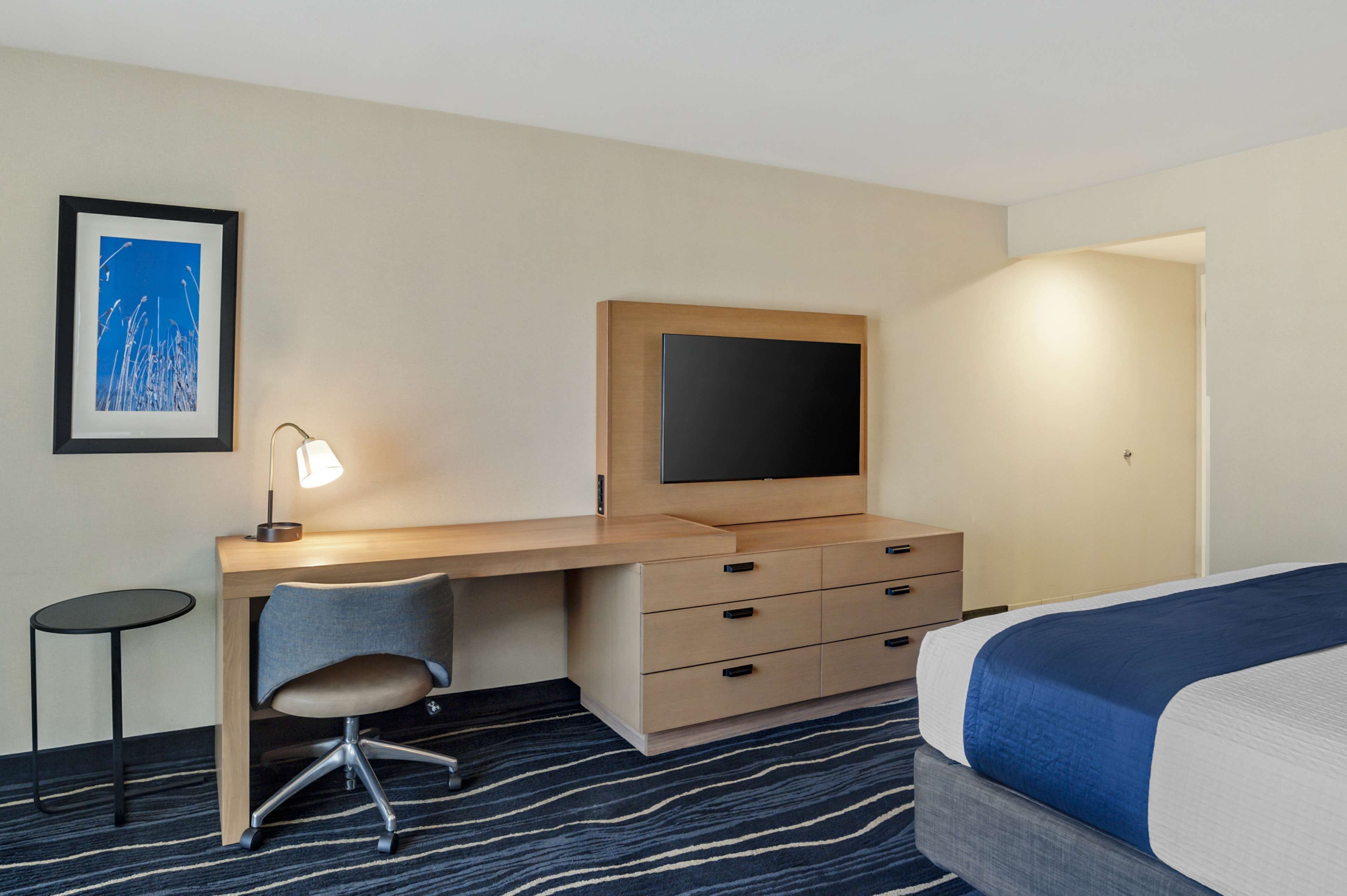Ett hotellrum med ett skrivbord, en stol, en tv, en säng och en inramad tavla på väggen.