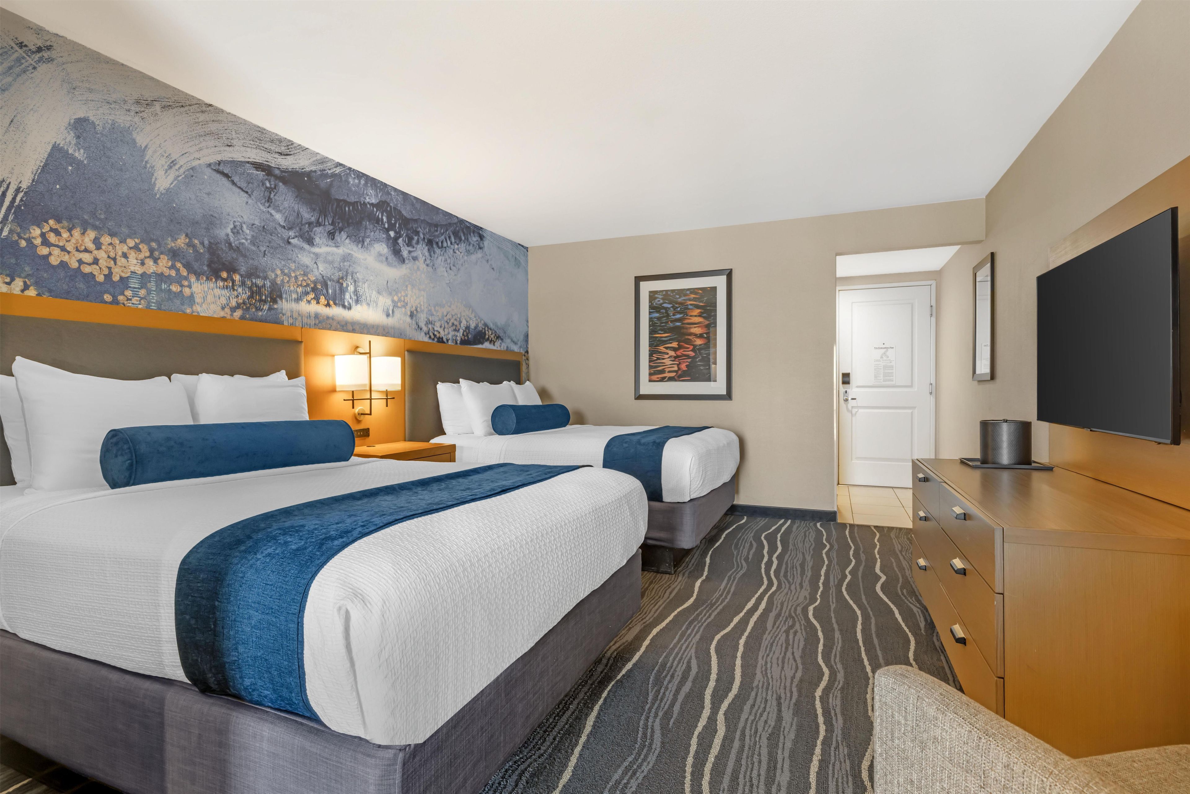 A hotel room with two beds, a large abstract painting on the wall, a flat-screen TV, and a wooden dresser.