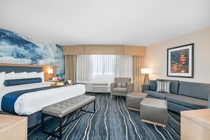 A hotel room with a large bed, a sofa, a chair, and a bench. There is a painting on the wall and a window with curtains.
