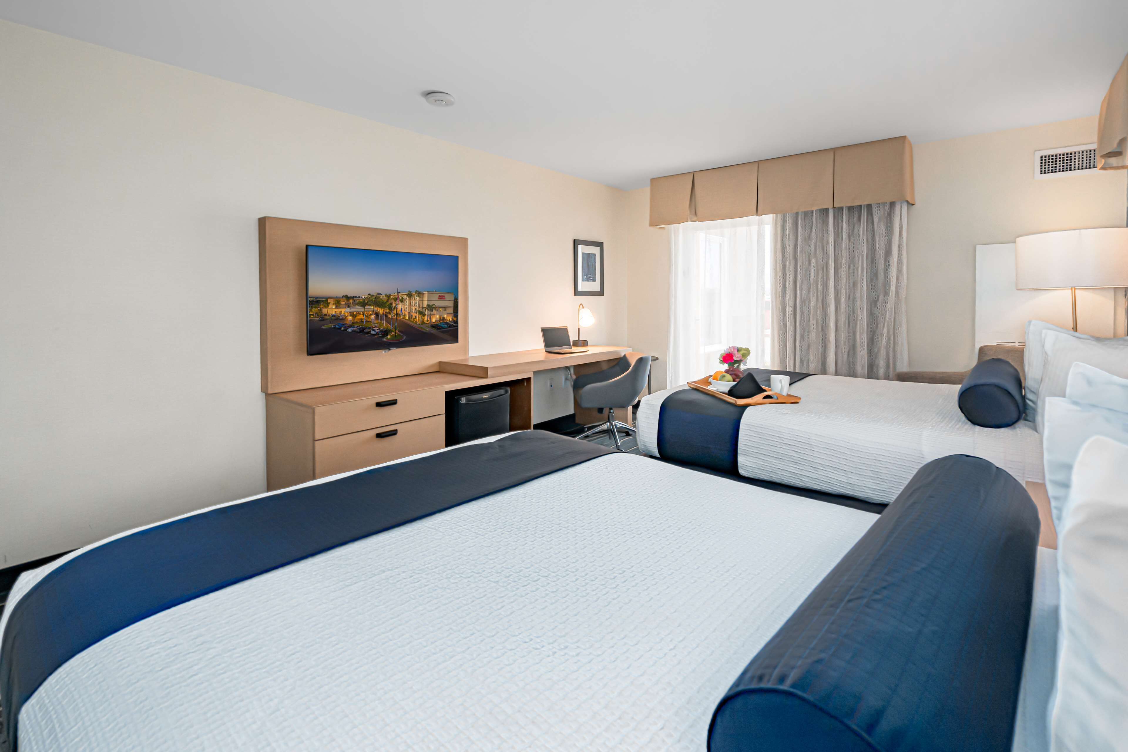 Une chambre d’hôtel avec deux lits, un bureau, une chaise, une lampe et une fenêtre avec des rideaux.
