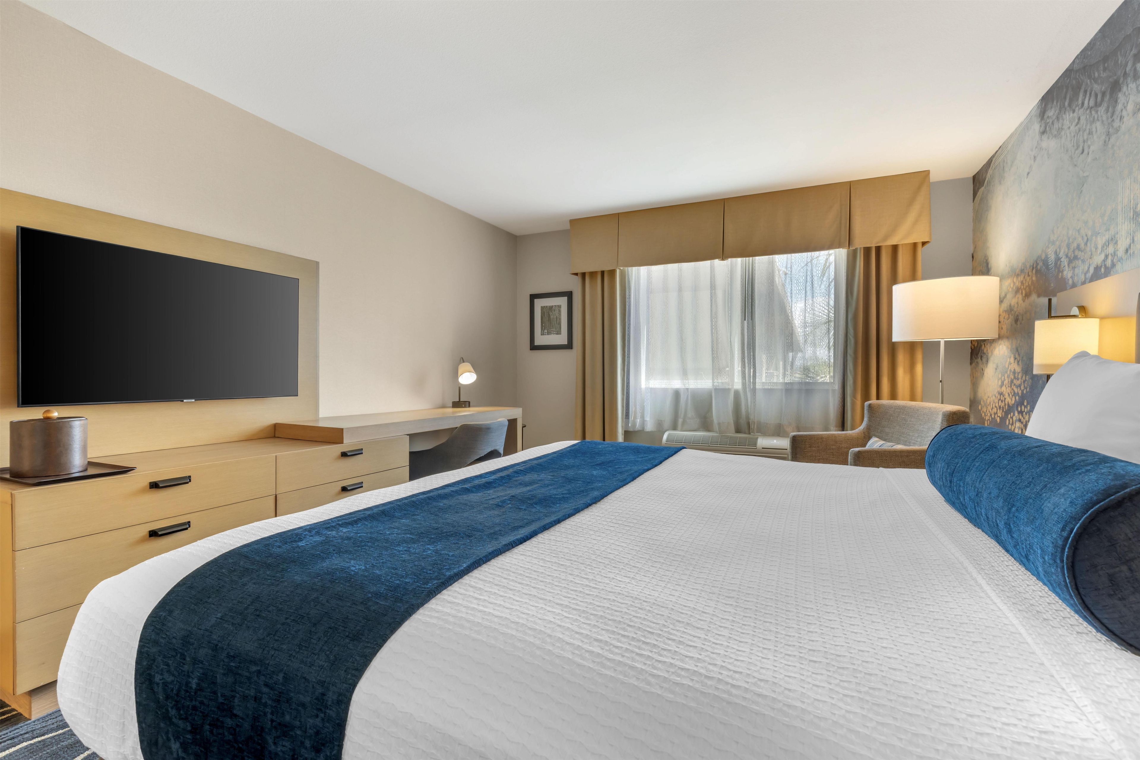 A hotel room with a large bed, a desk, a chair, a television, and a window with curtains.