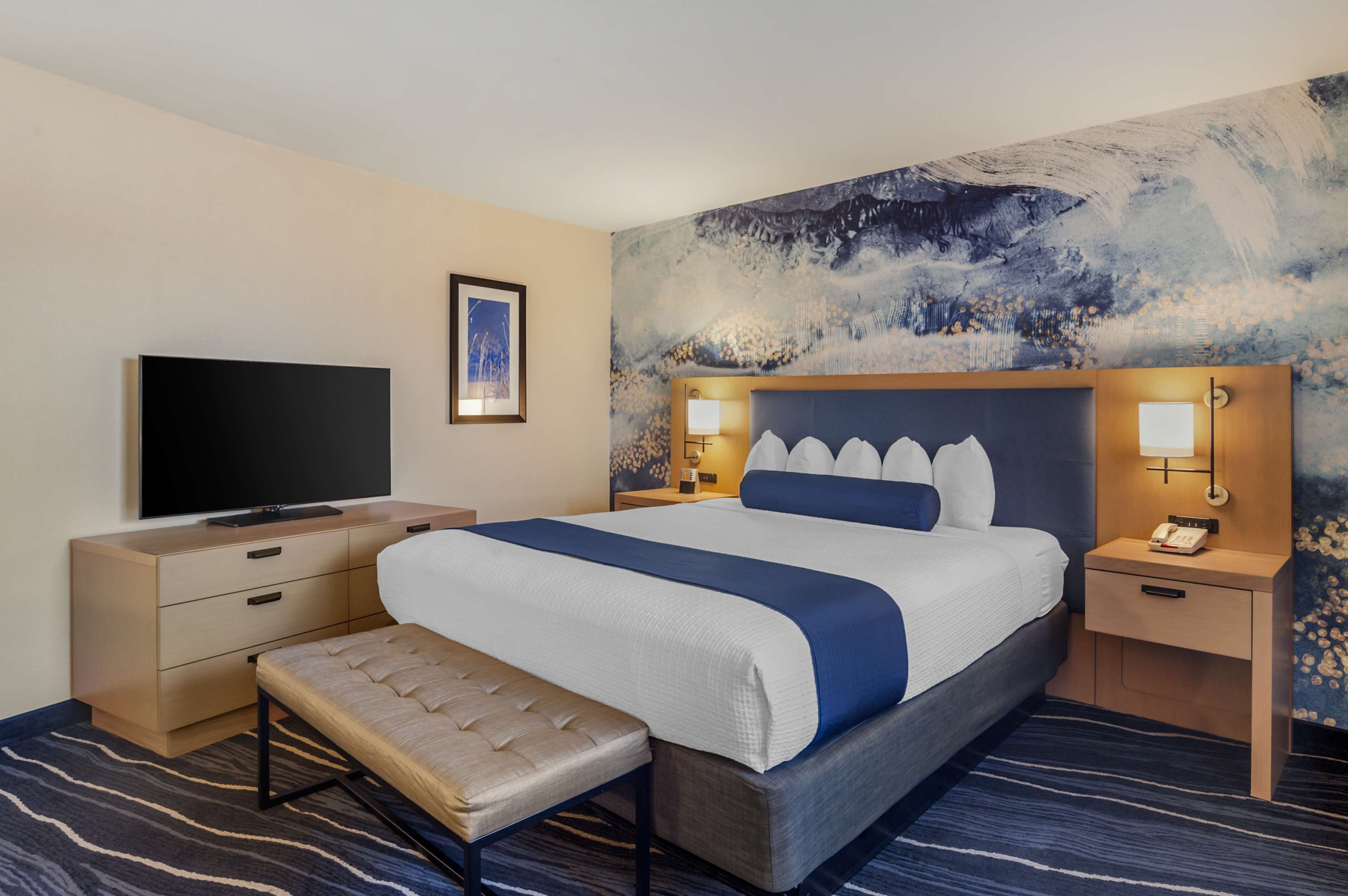 A hotel room with a large bed, a flat-screen TV, bedside lamps, a bench, and a wall mural.