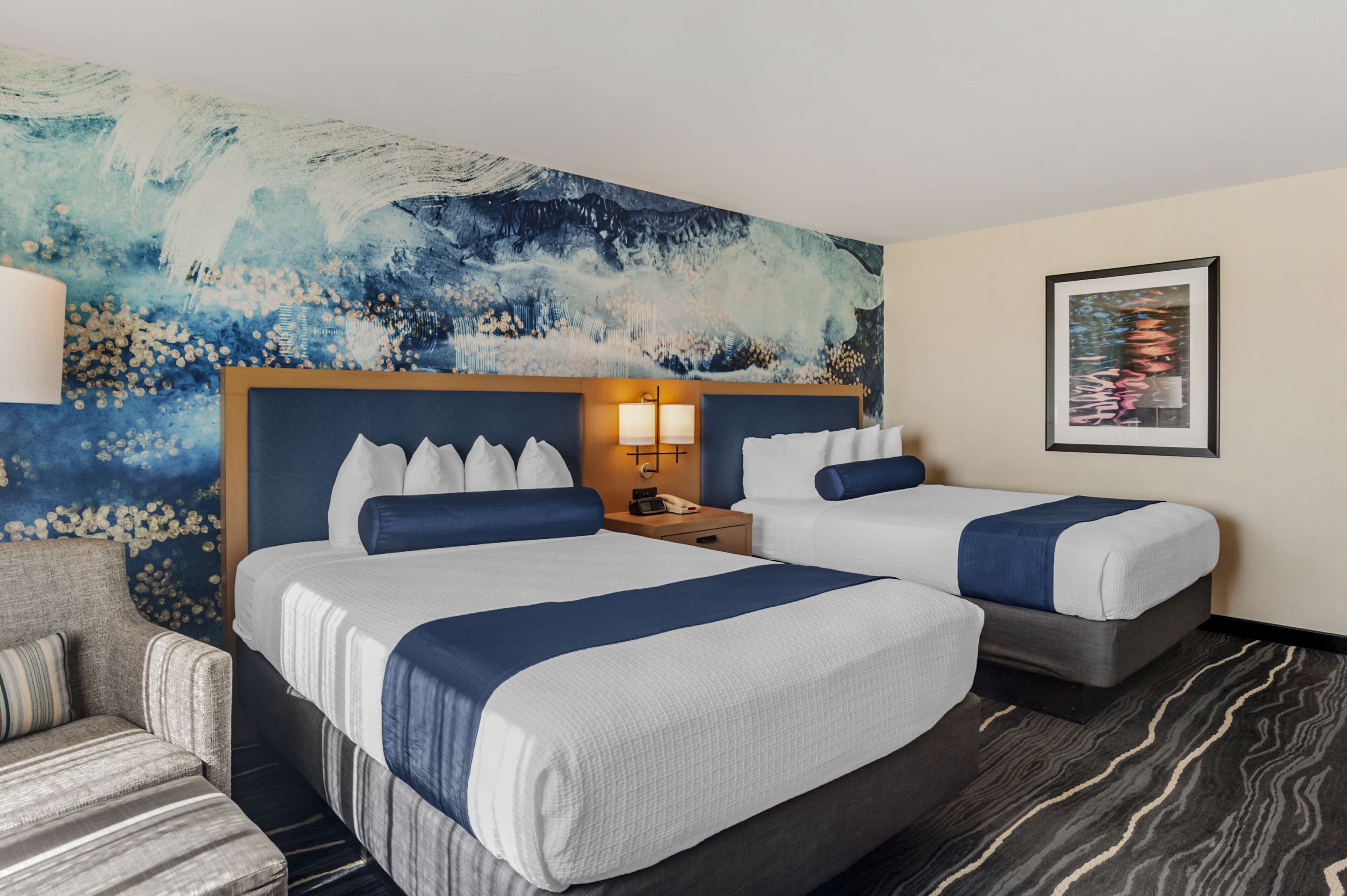 Habitación de hotel con dos camas, una mesita de noche, una lámpara, una silla y un cuadro abstracto grande en la pared.