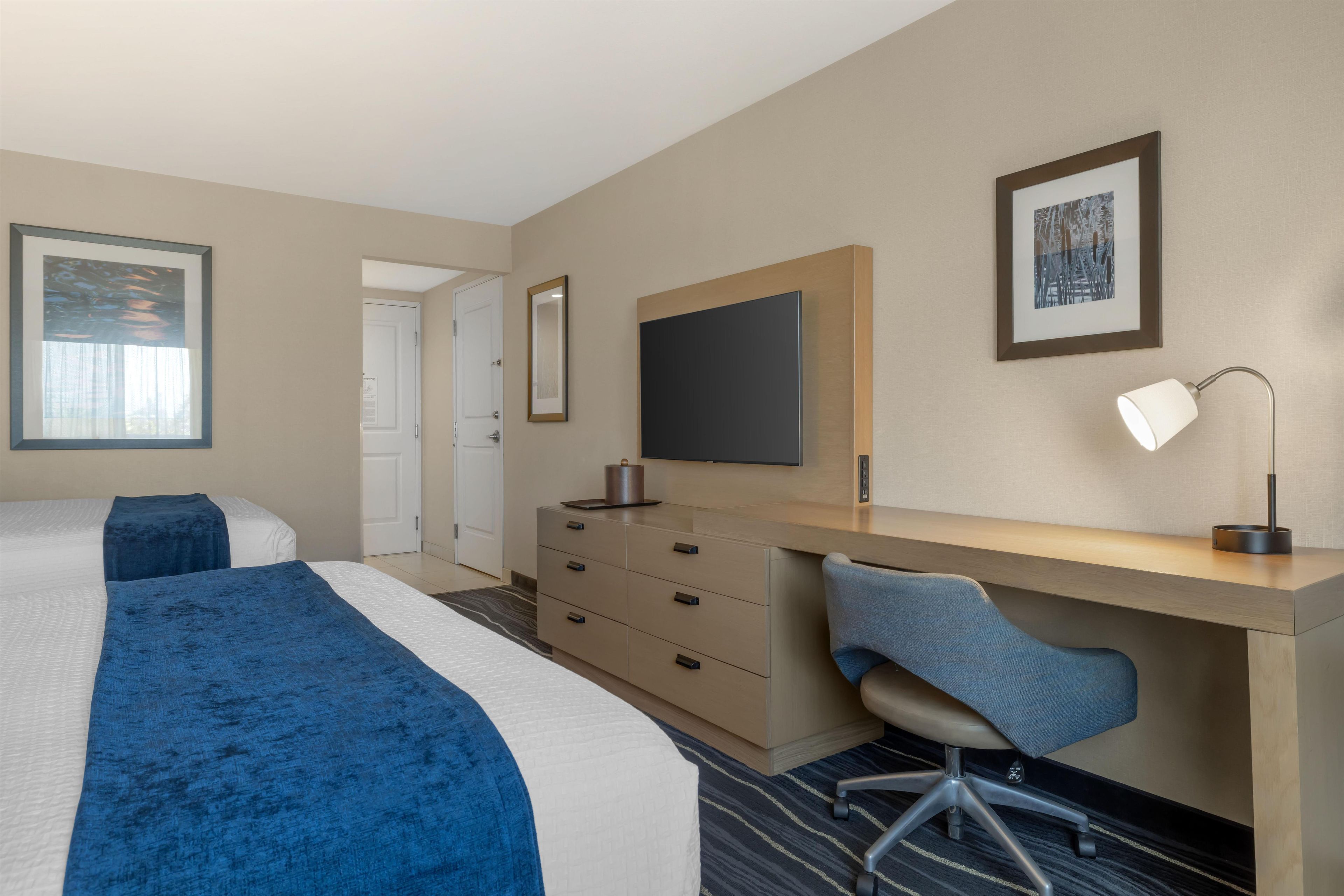 A hotel room with two beds, a desk, a chair, a flat-screen TV, and a lamp.