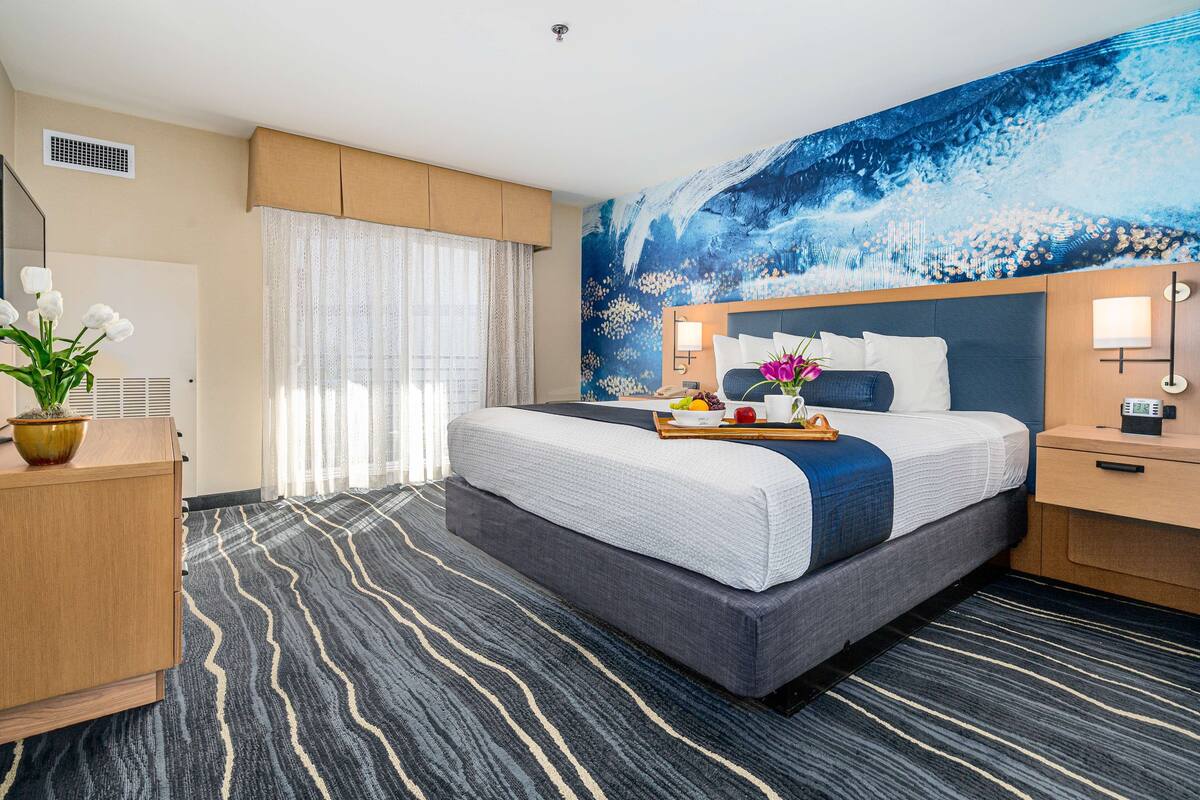 Habitación de hotel con una cama grande, un aparador de madera, un escritorio y un amplio ventanal con cortinas translúcidas.
