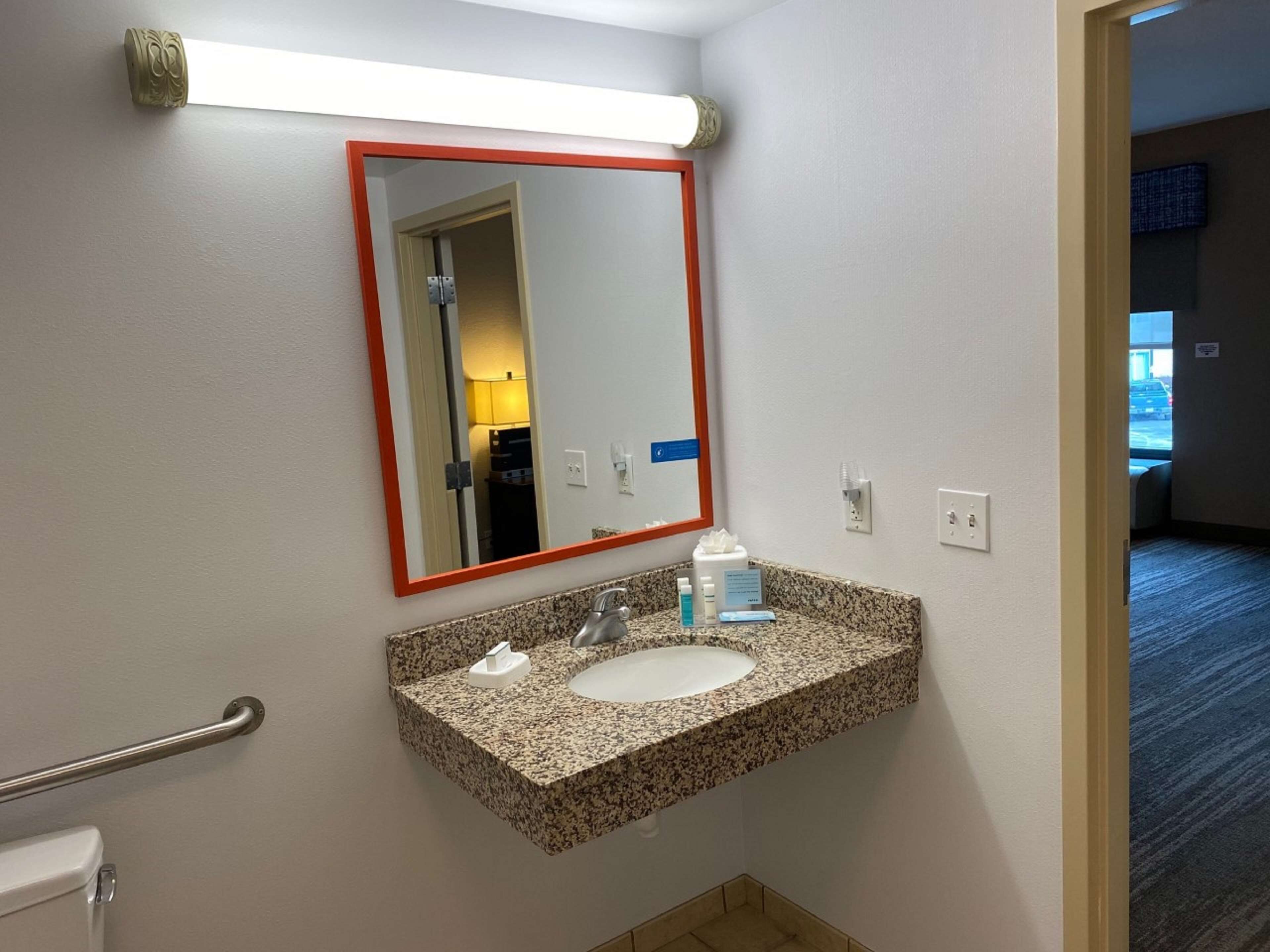 Une salle de bain avec un lavabo doté d’un plan de travail en granit, un miroir avec un cadre rouge et une barre d’appui.