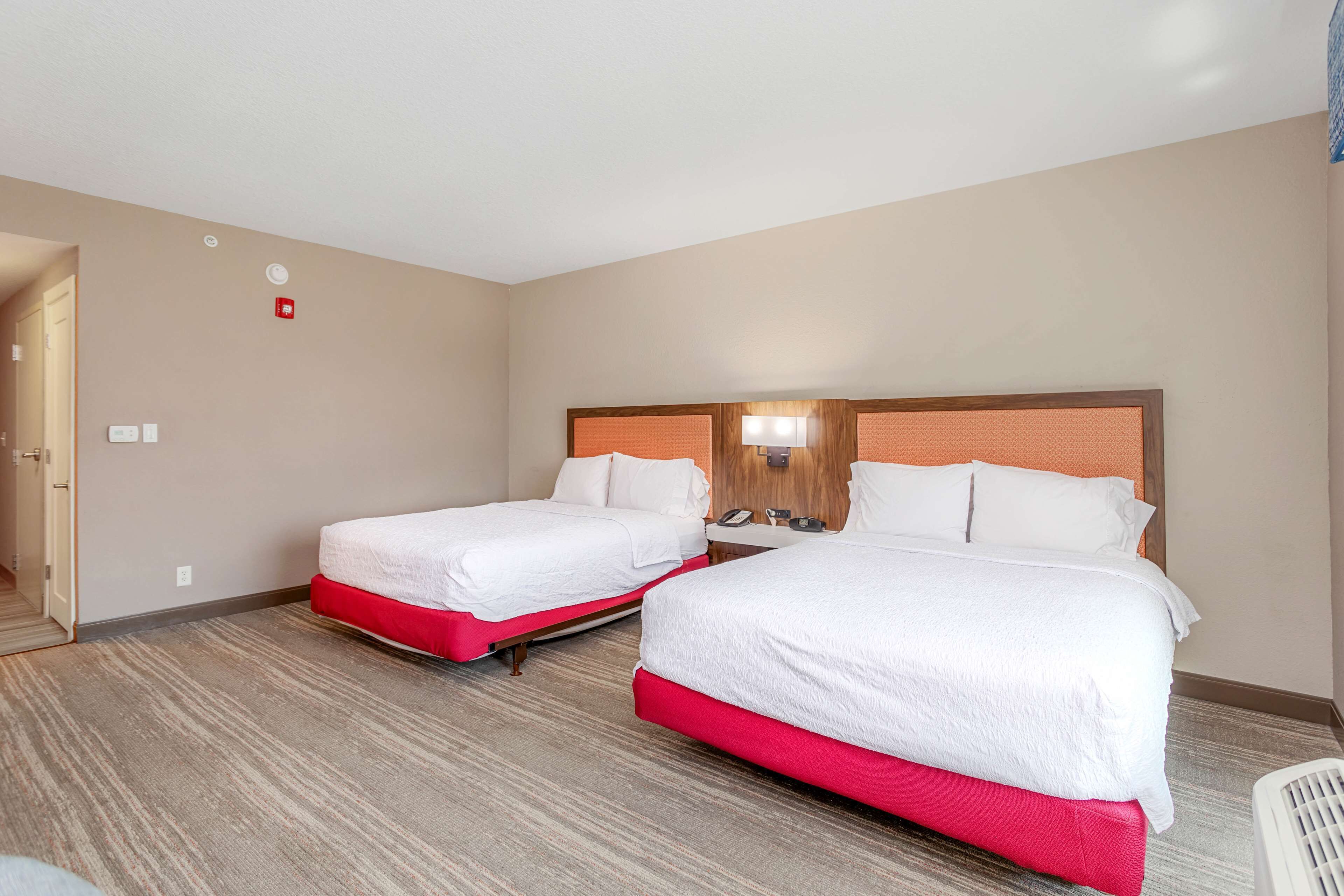 A hotel room with two beds, a wooden headboard, a bedside table with a lamp, and a red carpeted floor.