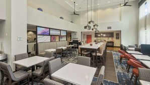 Un'ampia hall dell'hotel con diverse zone salotto, tra cui un lungo tavolo condiviso e numerosi posti a sedere riservati. Sono presenti ventilatori a soffitto, un angolo caffè e una zona cucina.