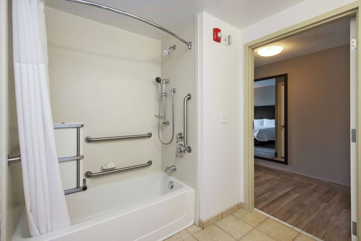 A bathroom with a bathtub, shower, handrails, and a door leading to a bedroom.