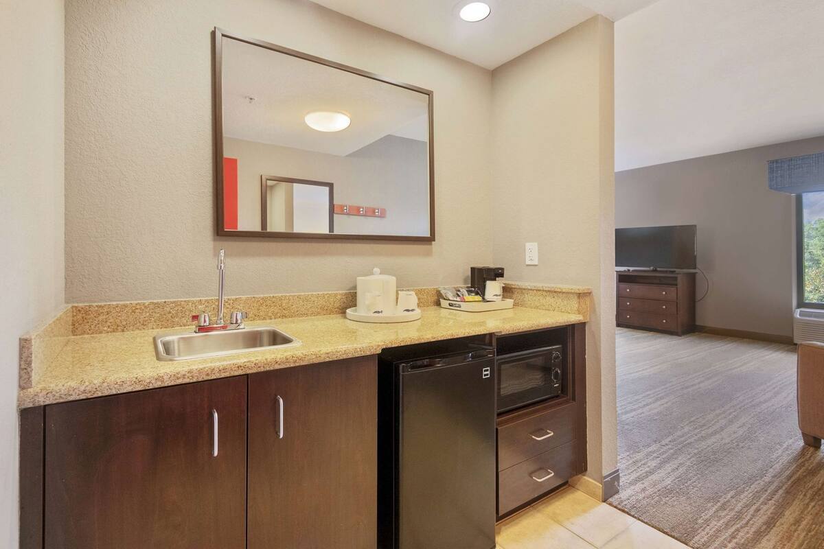 A compact kitchen with a sink, a coffee maker, and a microwave. There is a large mirror above the sink and a television in the adjacent room.