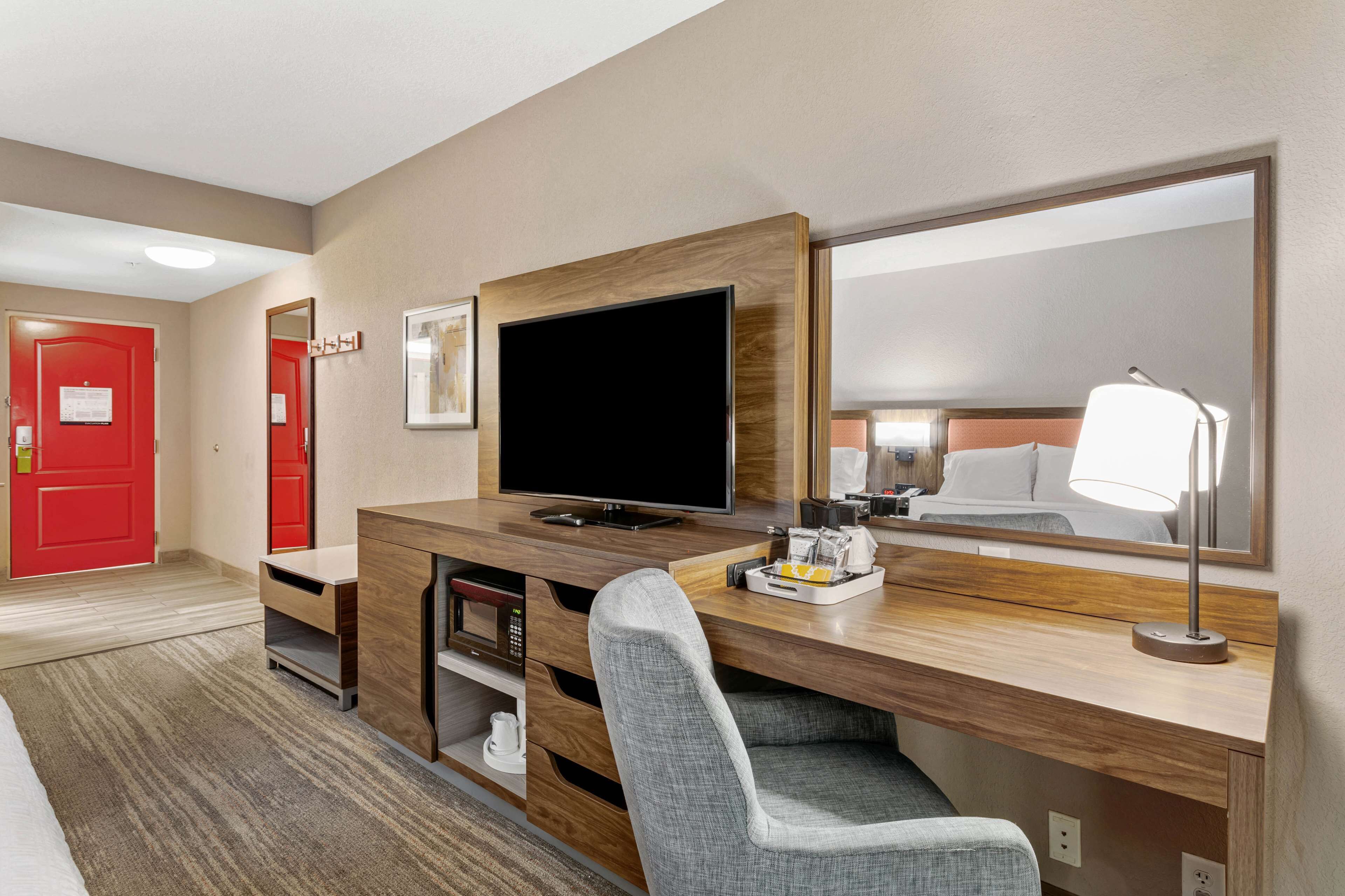 Ein Hotelzimmer mit einem Holztisch, einem Fernseher, einem Spiegel, einer roten Tür und einem Bett mit weißer Bettwäsche.