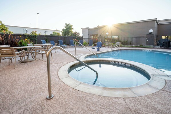 A swimming pool with lounge chairs and a sunshade, surrounded by a fence and a building in the background.