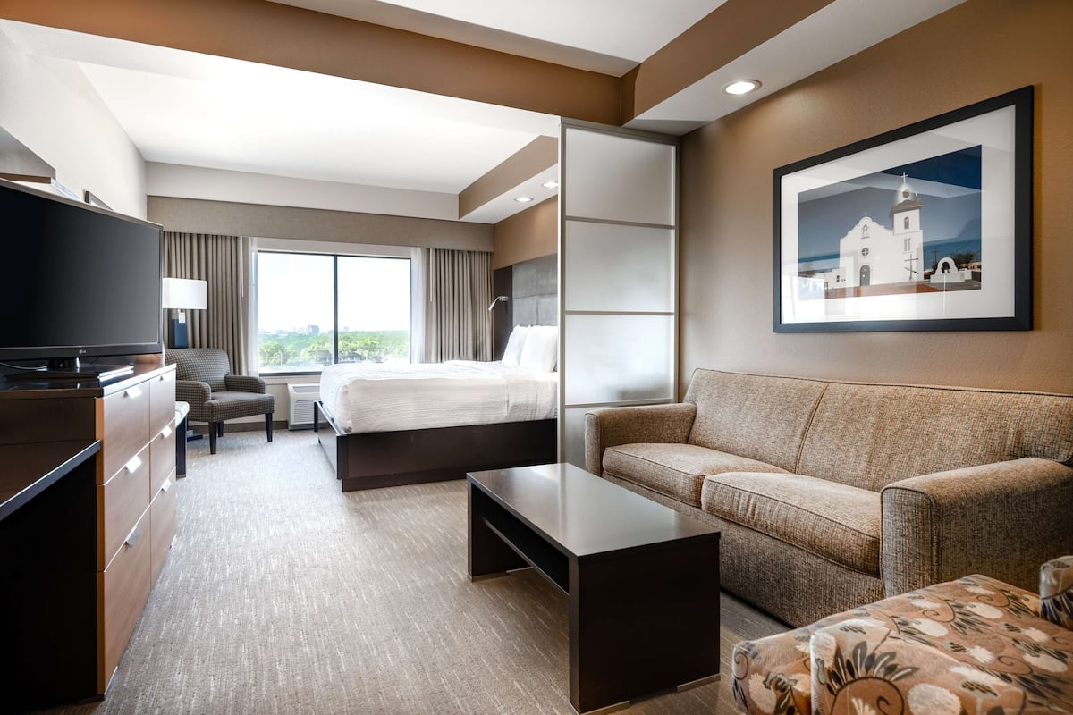 Ein modernes Hotelzimmer mit Bett, Sofa, Fernseher, Schreibtisch und einem großen Fenster mit Blick auf die Stadt.
