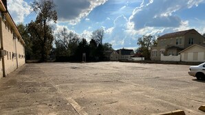 An empty parking lot with a building on the left and houses in the background.