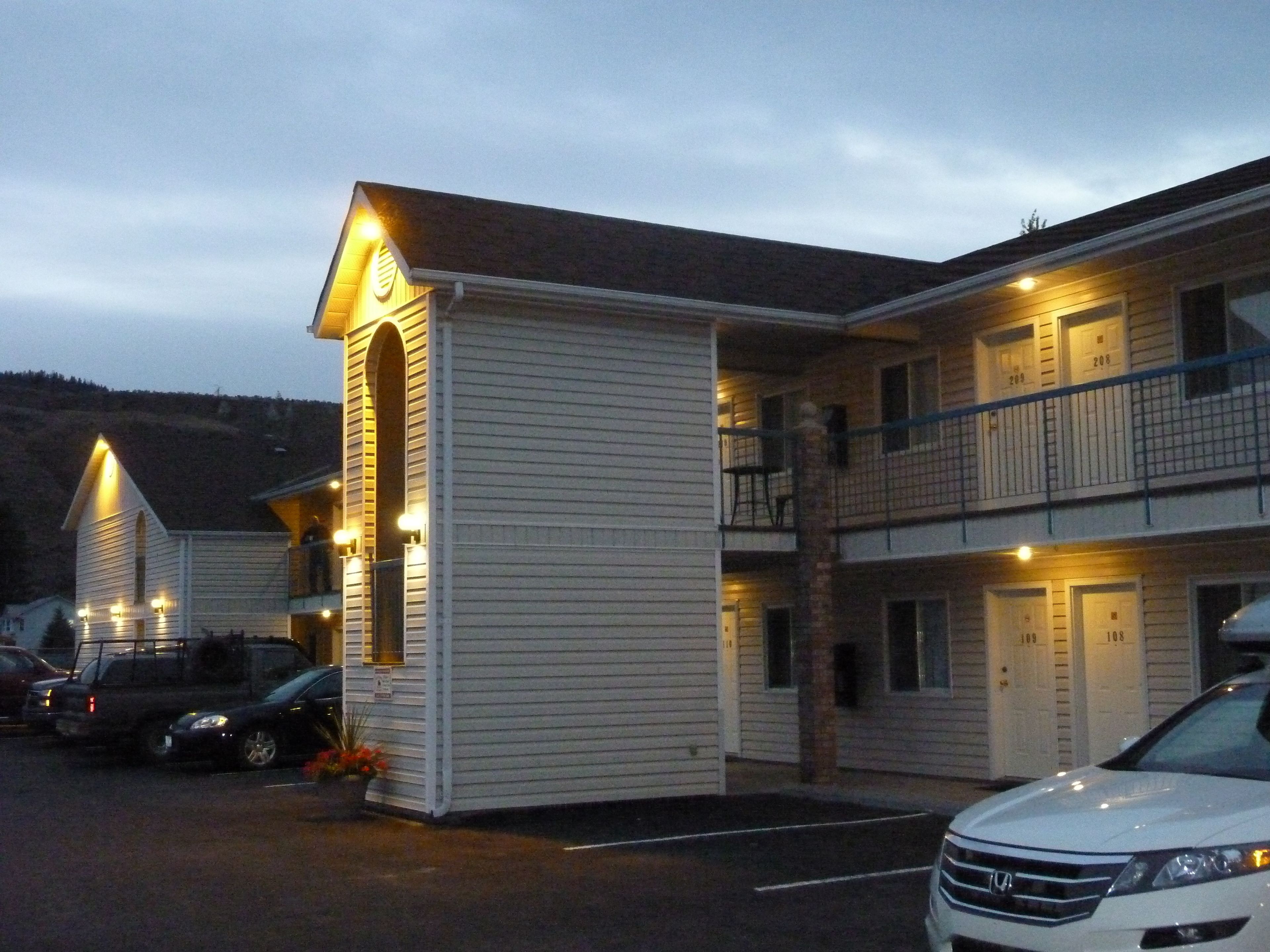 Un motel con una estructura de dos pisos, una zona de estacionamiento techada y un pequeño edificio adosado con techo inclinado.
