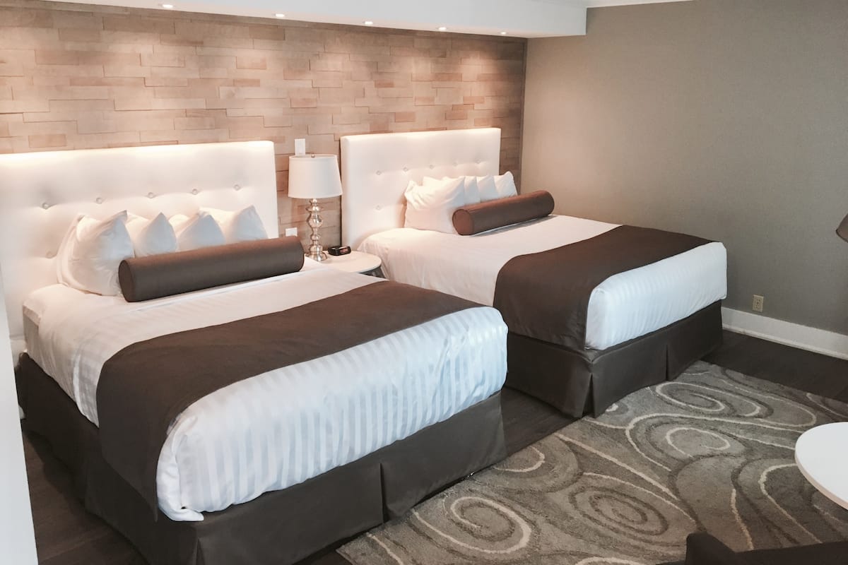 A hotel room with two beds, a headboard with a light fixture, a small table with a lamp, and a patterned rug.