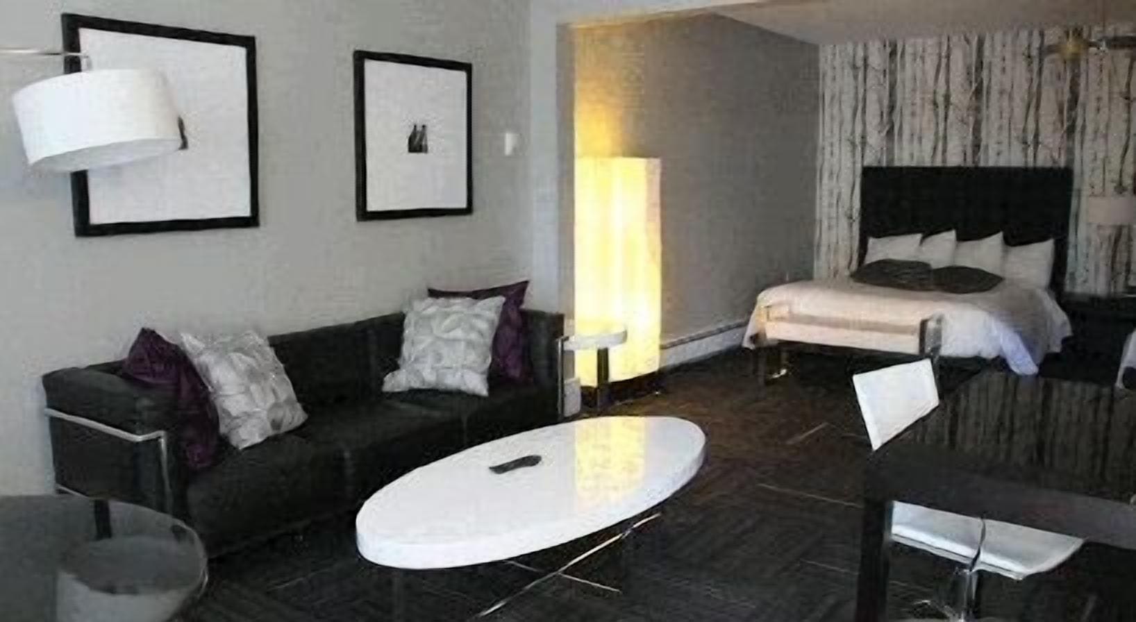 A modern hotel room with a black sofa, a round coffee table, a bed with a white headboard, and a large window with curtains.