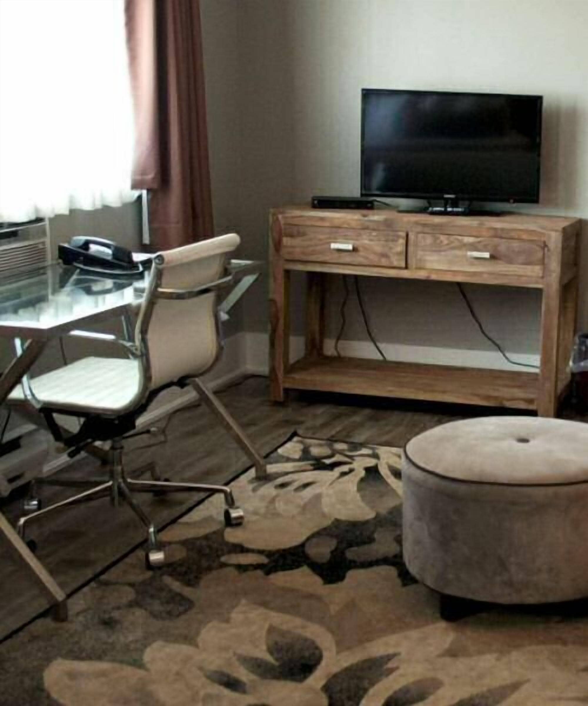 A hotel room with a glass desk, a chair, a television on a wooden stand, and a patterned rug.