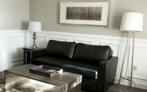 A living room with a black leather sofa, a patterned coffee table, a framed artwork on the wall, and a floor lamp. - Willow Bend Motel (Truro)
