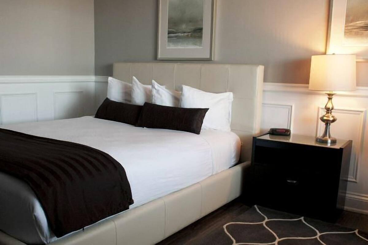 A neatly made bed with white and dark pillows, a bedside table with a lamp, and a framed picture on the wall.
