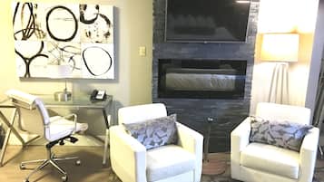 Una sala de estar moderna con chimenea de piedra, un televisor de pantalla plana montado encima, dos sillones blancos con cojines estampados, un escritorio de vidrio con un teléfono y una lámpara a la derecha.