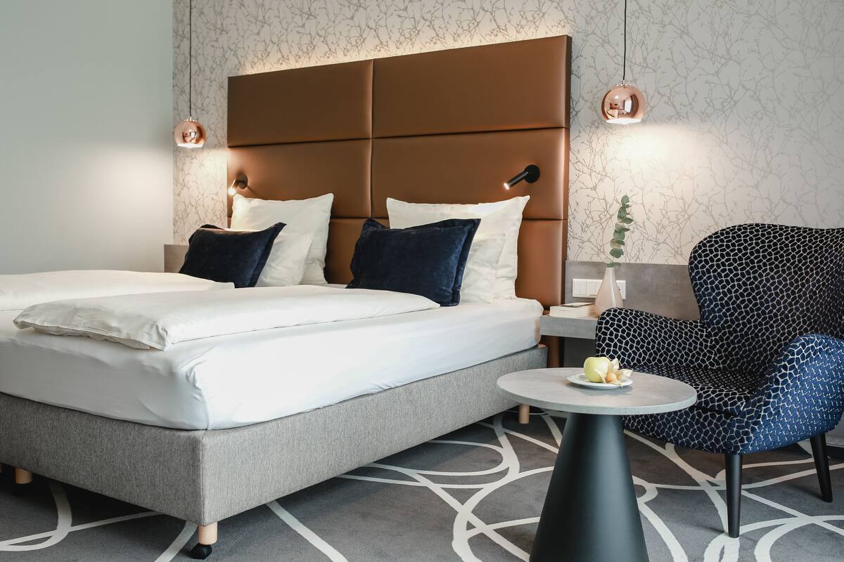 Una habitación de hotel moderna con una cama grande, una mesita redonda, un sillón estampado y una planta decorativa.