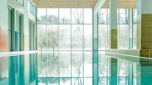 Una gran piscina interior con paredes de vidrio transparente, suelo de baldosas y vistas a árboles.