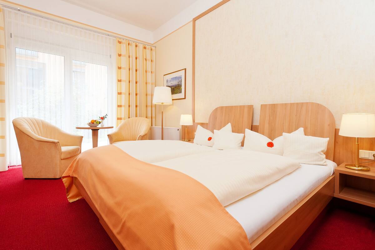 Habitación de hotel con una cama grande, dos mesitas de noche, una silla, una pequeña mesa con fruta y una ventana con cortinas.