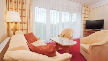 A hotel room with a sofa, armchair, small table, lamp, TV, and curtains.