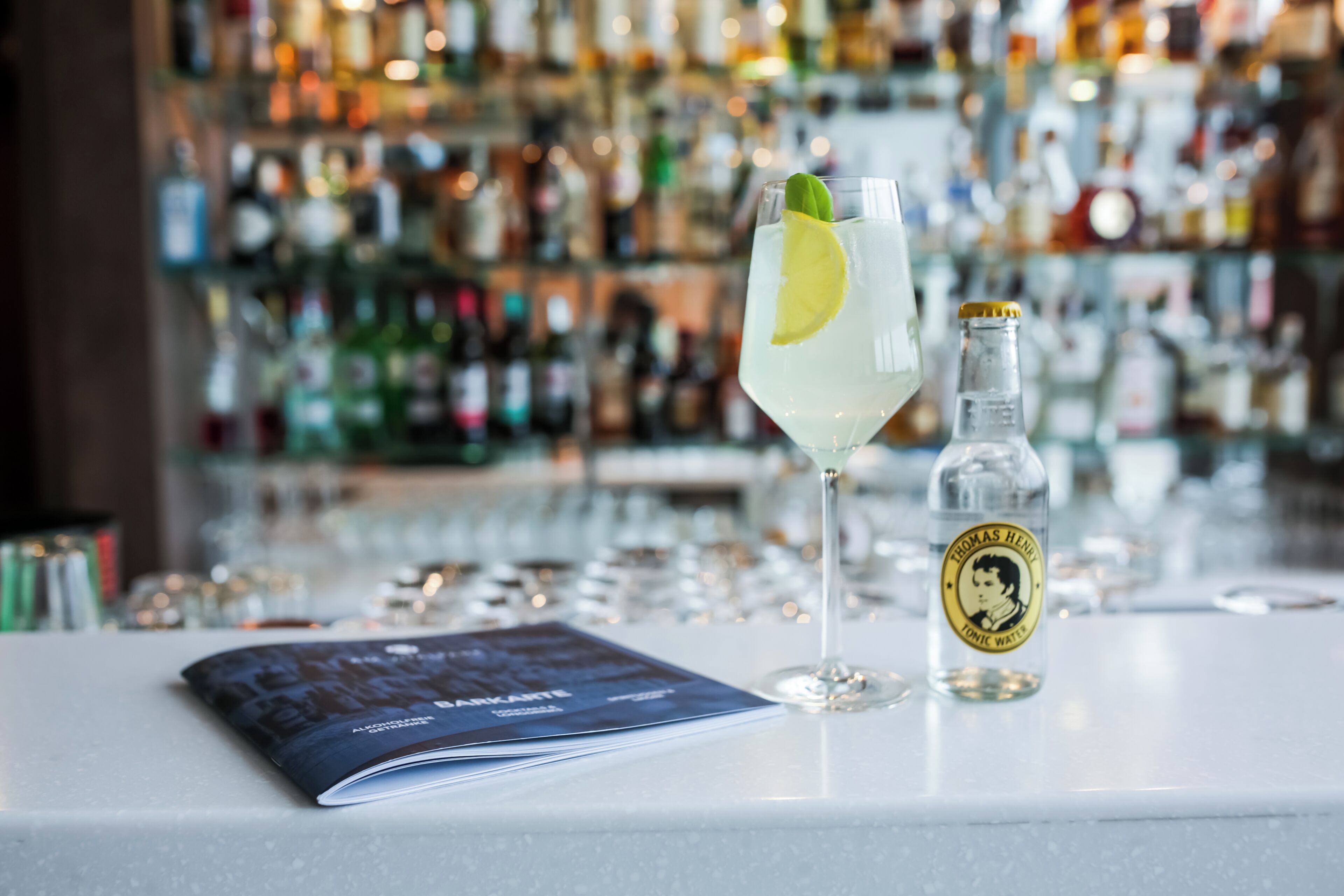 A bar setting with a glass of a clear beverage, a bottle of Thomas Hine Iodic Water, and a menu on the counter.