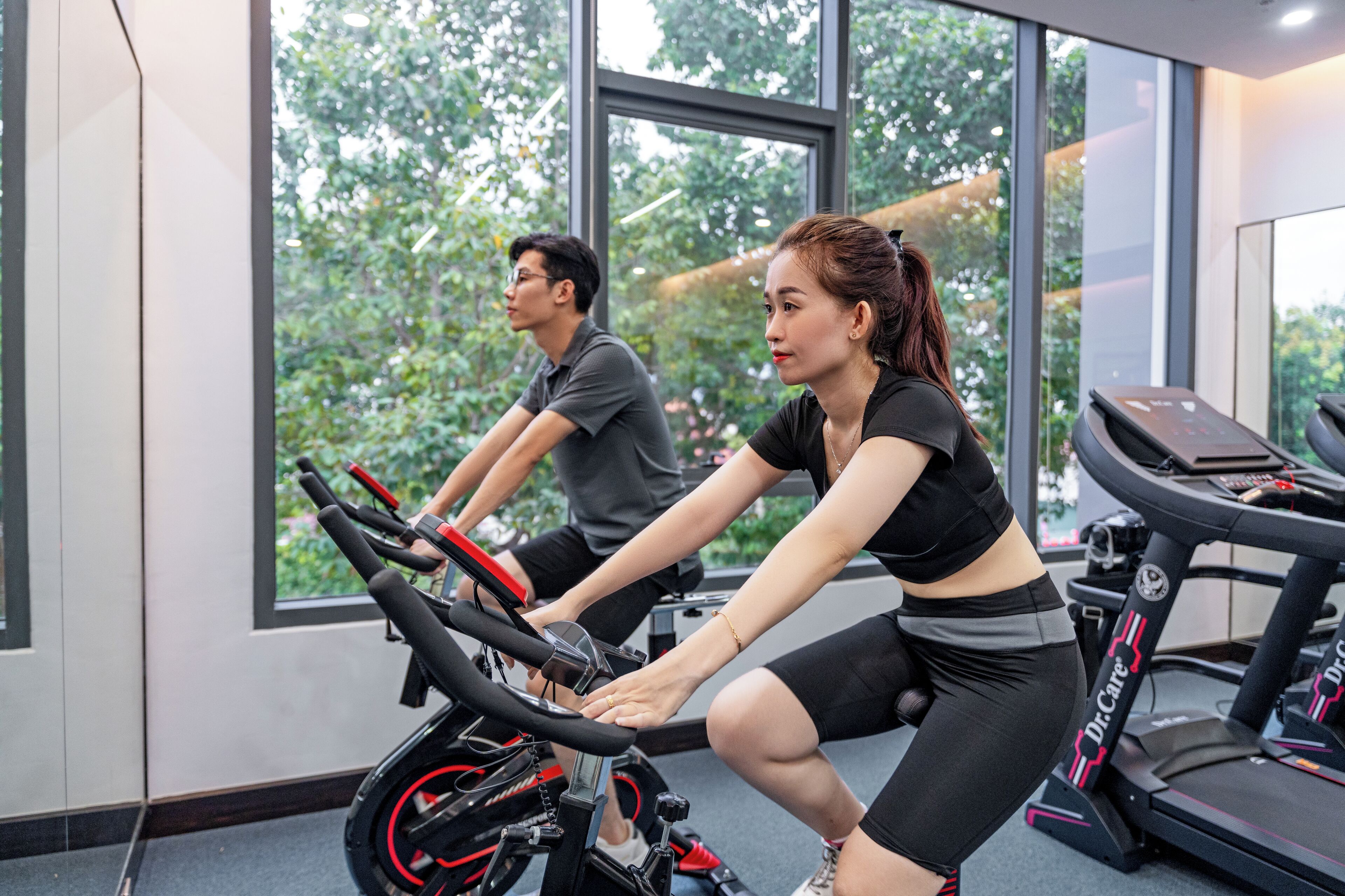 A gym with stationary bikes and treadmills, large windows with a view of trees, and a modern interior design.