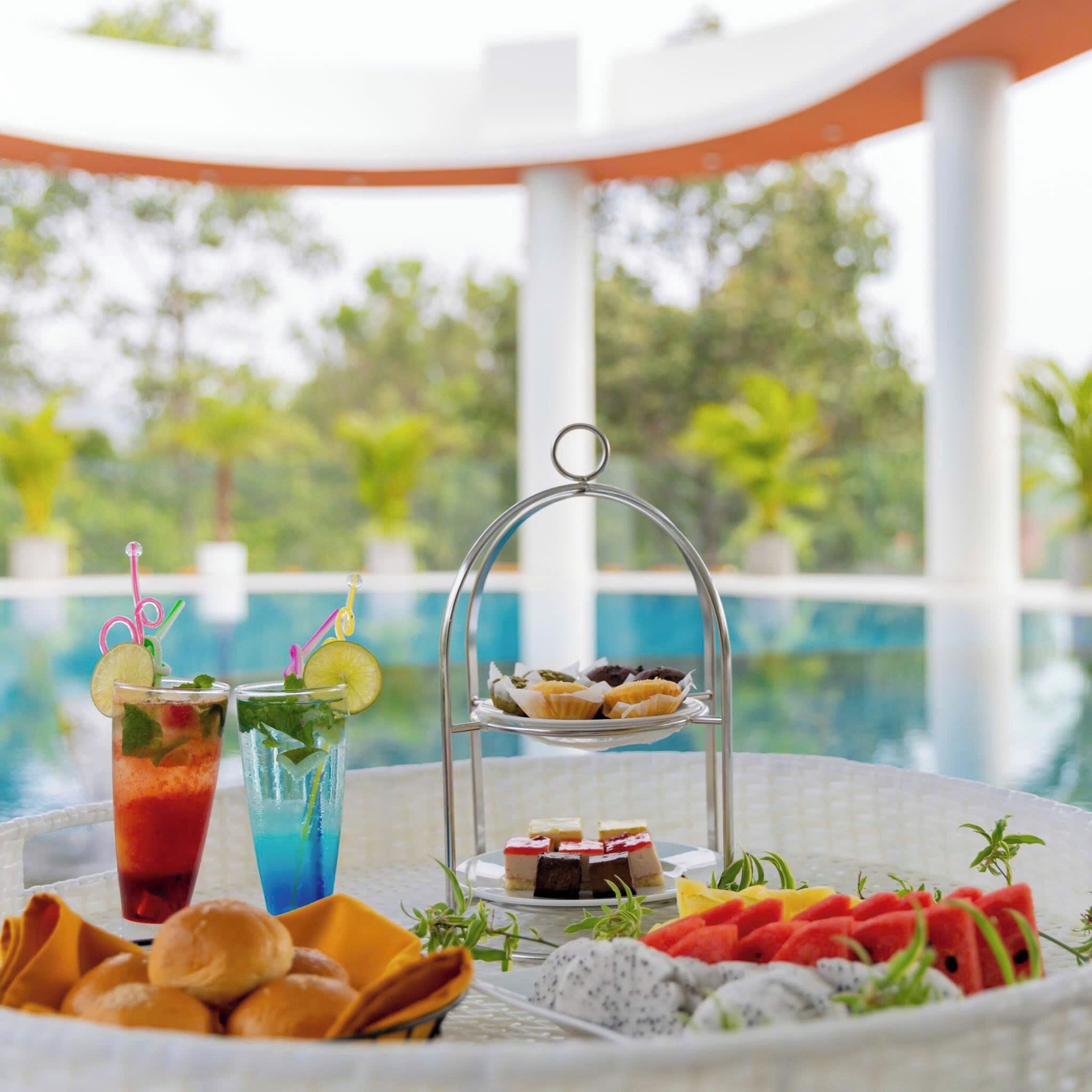 Une table garnie d’une variété de plats et de boissons, dont un plateau de pâtisseries et un verre agrémenté d’une tranche de citron vert, le tout avec une vue sur la piscine en arrière-plan.