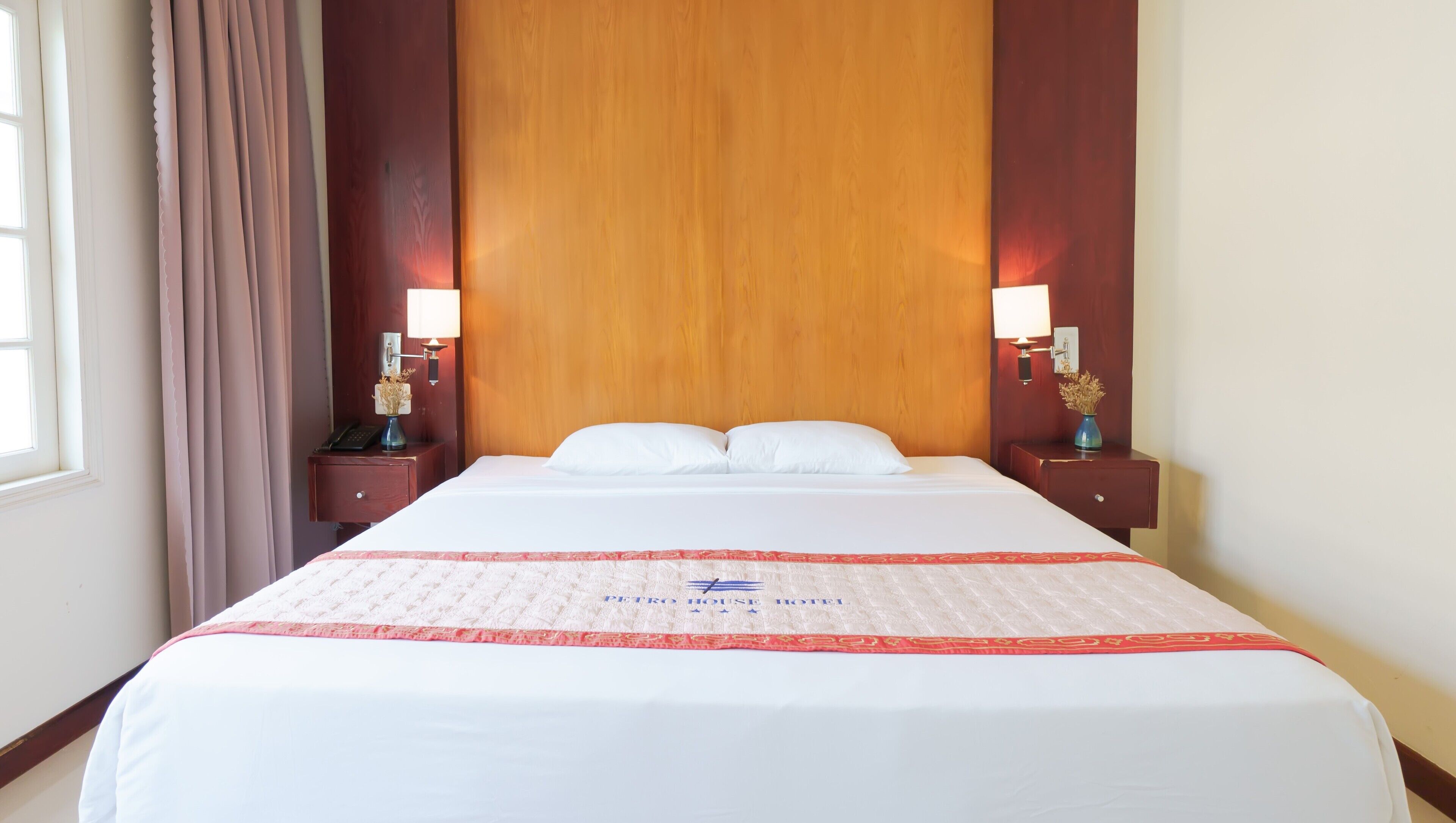 A hotel room with a large bed, two bedside tables with lamps, a wooden headboard, and a window with curtains.