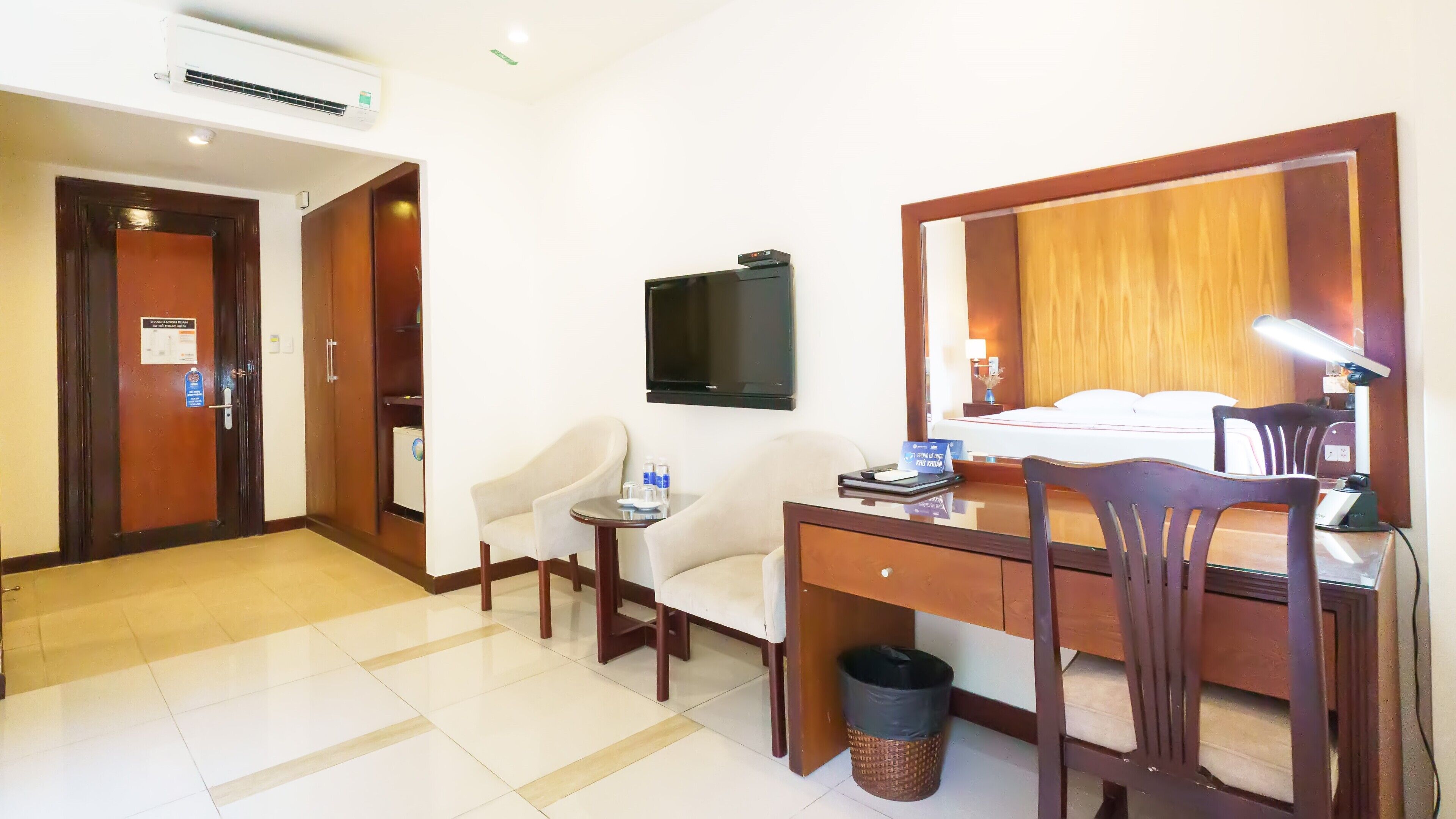A hotel room with a desk, two chairs, a television, a bed, and a mirror.