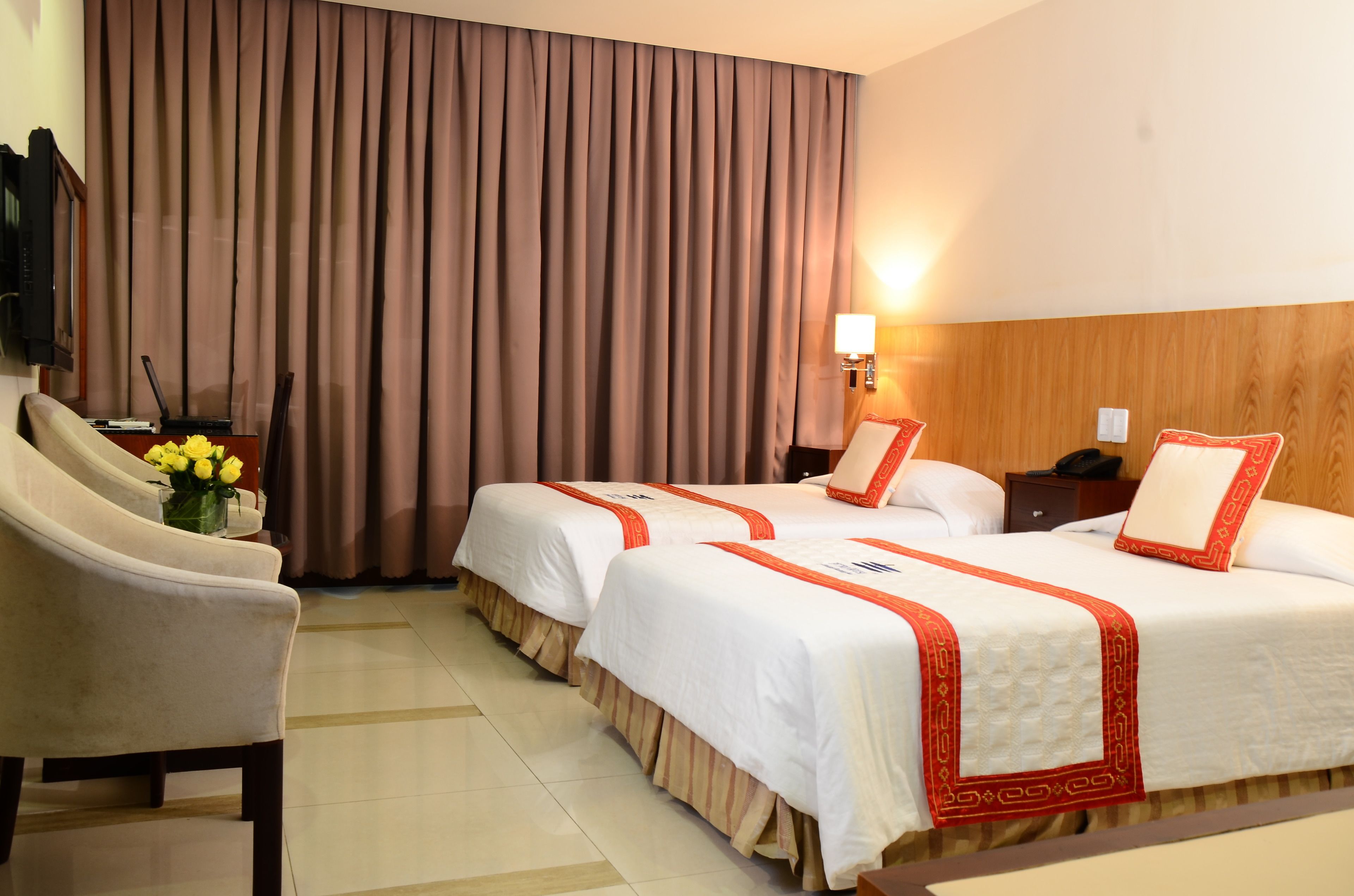 A hotel room with two beds, a desk, a chair, a lamp, and a large window with curtains.